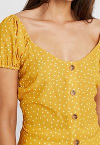 Donna che indossa un vestito giallo a maniche corte con pois, bottoni frontali e scollatura quadrata.