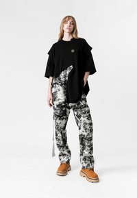 Mujer de pie vistiendo una camiseta negra oversized, pantalones abstractos en blanco y negro, y zapatos gruesos beige sobre fondo blanco.