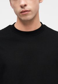 Νεαρός άνδρας με ανοιχτόχρωμο δέρμα, φορώντας απλό μαύρο μπλουζάκι με στρογγυλή λαιμόκοψη, φαίνεται από τον λαιμό μέχρι το πάνω μέρος του στήθους, με λευκό φόντο.