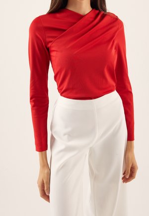 Femme portant un haut rouge à manches longues avec un décolleté asymétrique et un pantalon blanc taille haute, debout devant un fond uni.
