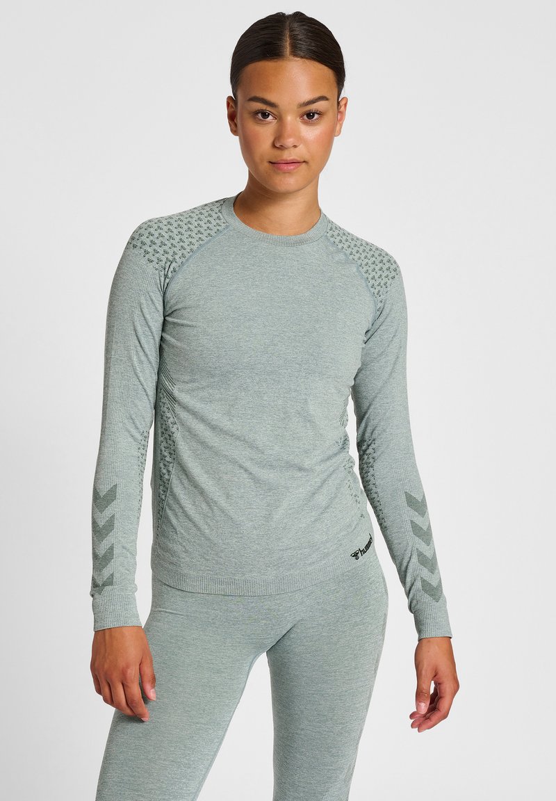 Jeune femme debout, face à nous, portant un haut à manches longues ajusté gris clair et un legging assorti avec des détails subtils de motifs.