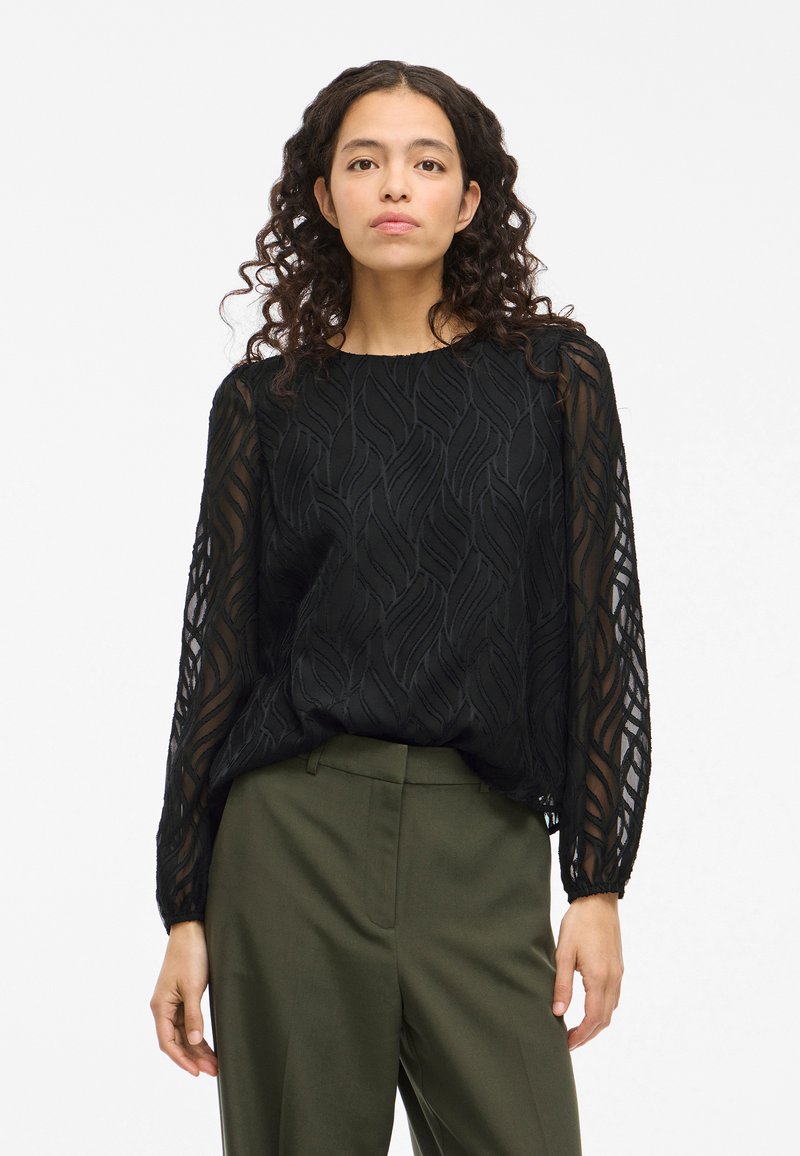 Black layered blouse featuring a leaf pattern, sheer long sleeves, and a rounded neckline, paired with dark green trousers.