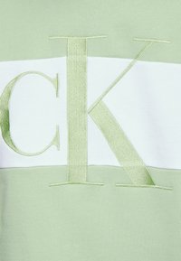 Sudadera verde menta con una franja horizontal blanca y un gran bordado en verde claro de las letras "CK". Textura suave y diseño casual.