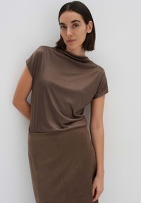 Bruine top met korte mouwen en een gedrapeerde halslijn, gecombineerd met een bijpassende bruine rok. Gladde stof met een zachte textuur en minimale ontwerpdetails.