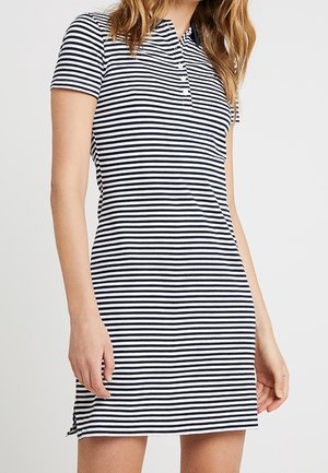 Woman wearing short-sleeve black and white horizontal striped polo dress with buttoned collar, standing against plain light background.