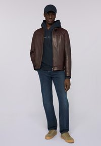 Chaqueta de cuero marrón con cremallera frontal y bolsillo lateral, sudadera azul marino debajo, vaqueros de denim azul y zapatillas color beige con detalle texturizado.
