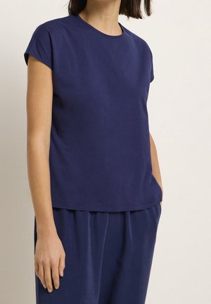 Kobieta ubrana w gładki, granatowy top z krótkim rękawem i dopasowane luźne spodnie, stojąca z jedną ręką w kieszeni na neutralnym tle.