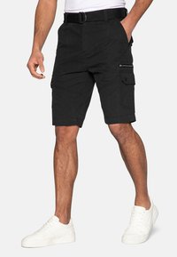 Shorts cargo noirs en tissu résistant, avec des poches latérales, une ceinture et une coupe à hauteur des genoux. Associés à des baskets blanches.