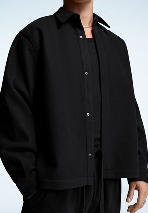 Personne portant une chemise noire ample boutonnée sur un t-shirt noir, un pantalon noir assorti, un collier en chaîne et une bague, la main dans la poche.
