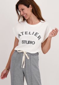 Witte t-shirt met korte mouwen met zwarte tekst "ATELIER" en "STUDIO," gecombineerd met grijze joggingsbroek met trekkoord. Soepele stof en comfortabele pasvorm.