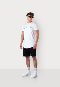 Weißes Baumwoll-T-Shirt mit "STLIM"-Schriftzug, schwarze Shorts, weiße Turnschuhe und sportliche Sonnenbrille. Flaches Design, kurze Ärmel, lockerer Schnitt.