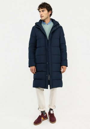 Hombre con un abrigo acolchado largo azul marino, suéter azul claro, pantalones blancos y zapatillas burdeos, de pie contra un fondo liso mirando hacia la izquierda.
