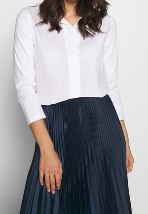 Vrouw draagt een witte blouse met lange mouwen, gestopt in een hoge taille, donkerblauwe geplooide rok, staande tegen een effen achtergrond.
