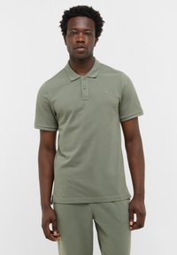 Mężczyzna stojący w oliwkowej koszulce polo z krótkim rękawem i dopasowanych spodniach na jednolitym białym tle.