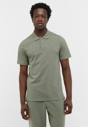 Mann, der ein olivgrünes Kurzarm-Polohemd und passende Hose vor einem schlichten weißen Hintergrund trägt.