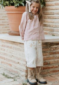 Blusa a cuadros en tonos rosas con mangas largas, pantalones blancos bordados y botas beige con cordones, de pie cerca de una maceta sobre un fondo de pared de piedra.
