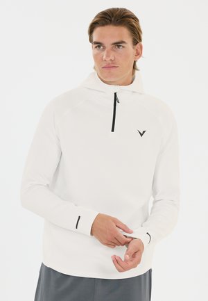 Jeune homme ajustant la manche d'un sweat à capuche blanc avec une fermeture éclair noire et un petit logo noir sur la poitrine.