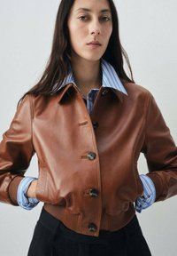 Veste bombardier en cuir marron avec une fermeture à boutons, des poignets et un col côtelés. Portée par-dessus une chemise rayée bleu et blanche.