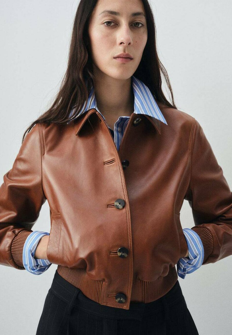Veste bombardier en cuir marron avec une fermeture à boutons, des poignets et un col côtelés. Portée par-dessus une chemise rayée bleu et blanche.