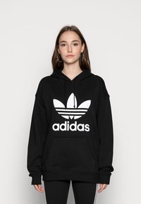 Juodas kapotas, pagamintas iš medvilnės, su dideliu baltu Adidas logotipu ir ikonine trijų juostelių dizainu priekyje. Įskaičiuota kengūros kišenė.