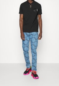 Schwarzes Poloshirt mit Logo-Patch, hellblaue Jeans mit wiederholtem Textmuster und schwarze Sneakers mit pinkfarbenen Akzenten und Schnürsenkeln.