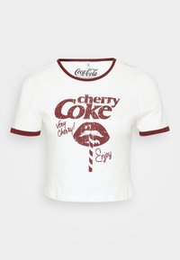 Bela skrajšana majica s kratkimi rokavi z okroglo češnjevo ovratnikom in obrobami rokavov. Ima retro rdeče grafično besedilo, ki pravi "Cherry Coke", in ilustracijo ust.