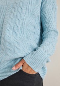 Jasnoniebieski sweter z dzianiny z wzorem warkocza, z prążkowanymi mankietami i dołem. Połączony z ciemnym materiałem, ukazującym teksturę i kontrast kolorów.