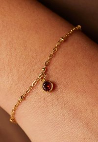 Gouden armband met een ronde rode edelsteen hanger. De ketting bestaat uit onderling verbonden schakels en ligt op een textuur huid.