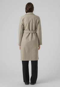 Beige wollen blend lange jas met een riem, voorzien van een reverskraag en lange mouwen, gedragen over zwarte broeken. Gladde textuur en knielang.