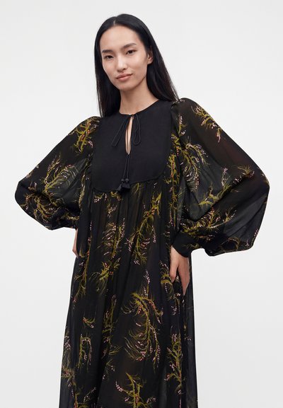 Femme portant une robe noire transparente avec des motifs floraux verts et roses et des manches bouffantes volumineuses, debout les mains dans les poches.