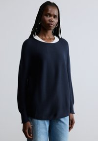 Laivastonsininen neulepaita, jossa pitkät hihat, pyöreä pääntie ja väljä istuvuus, yhdistetty vaaleansinisin farkkuihin. Sileä pinta ja rento siluetti.