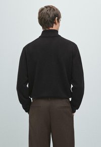 Polo in maglia nera con colletto alto, vestibilità rilassata e maniche lunghe, abbinato a pantaloni marroni con tasche posteriori. Design minimalista.