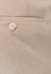 Beige szövet finom textúrával, egy kis, kerek gombbal és egyetlen zsebbel. Az élek mentén a varrás részletei láthatók.