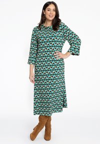 Grünes, gemustertes Kleid mit drei Viertel langen, ausgestellt geschnittenen Ärmeln, das geometrische Formen in Blau, Orange und Weiß zeigt. Kombiniert mit braunen, knielangen Stiefeln.