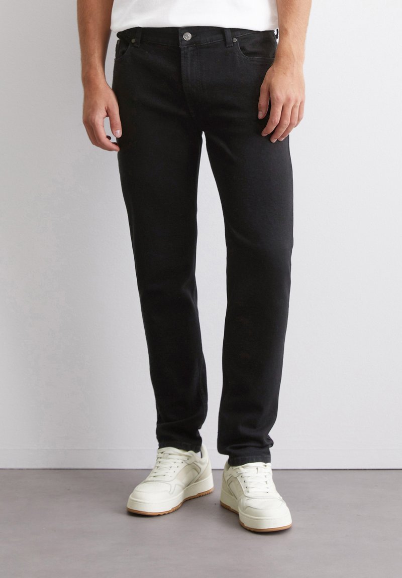 Fekete farmernadrág, slim fit szabással, gombzárral és öt zsebes kialakítással. Fehér sportcipővel és sima pólóval párosítva.