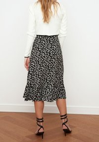 Sort midi-nederdel med blomsterprint og flæsekanter. Sammen med sorte højhælede sandaler med stropper. Baggrunden er helt hvid.