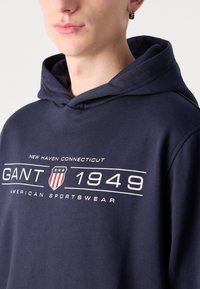 Tamnoplavi dukserica od pamuka s prednjim logotipom koji sadrži bijeli tekst i dizajn štita. Ima kapuljaču s vezicom.