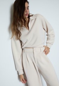 Frau in lockerer beiger Bluse und hochgeschnittenen beigen Hosen, die gegen eine weiße Wand steht und eine Hand in der Tasche hat.