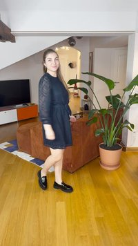 Fekete ruha áttetsző, hosszú ujjakkal és derékban megkötős résszel, fényes fekete loaferekkel párosítva. A háttérben egy cserepes növény és bútorok láthatók.