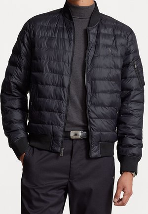 Mann trägt eine schwarze gesteppte Bomberjacke über einem dunkelgrauen Rollkragenpullover mit schwarzen Hosen und einem Gürtel mit silberner Schnalle.