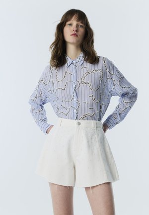 Vrouw die een blauw-wit gestreept blouse met knopen draagt met zwarte letters en witte shorts met hoge taille met kleine getextureerde stippen.
