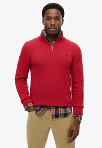 Jersey de punto rojo con cuello alto y cierre en el cuello, superpuesto a una camisa de cuadros. Presenta puños y dobladillo de canalé, tejido texturizado.