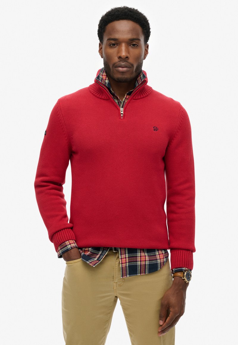 Jersey de punto rojo con cuello alto y cierre en el cuello, superpuesto a una camisa de cuadros. Presenta puños y dobladillo de canalé, tejido texturizado.