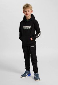 Svart sweatshirt med "hummel"-logotyp, matchande svarta byxor och blå träningsskor med vita detaljer och en texturerad sula. Svart hoodie med framficka.