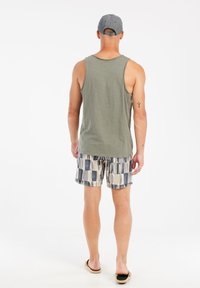 Casual outfit voor mannen: olijfgroen mouwloos shirt, gebloemde zwembaden in beige, marineblauw en lichtgrijs, zwarte sandalen en een grijze pet.