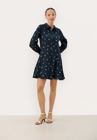 Robe chemise bleu marine avec manches longues, patte de boutonnage et motifs floraux blancs. Ourlet évasé, fabriquée dans un tissu lisse, associée à des talons roses.