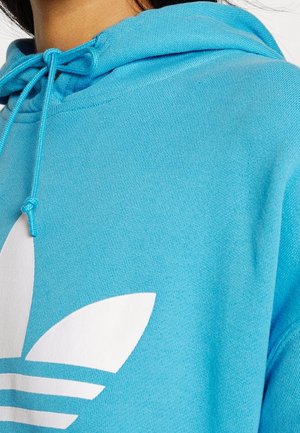 Gros plan d'une personne portant un sweat à capuche bleu vif avec un logo stylisé blanc à trois feuilles et des cordons attachés en nœud.