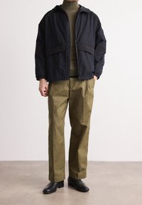 Černá bunda na zip s kapucí a kontrastní texturou. Olivově zelené kalhoty s volným střihem. Černé boty dokreslují vzhled.