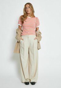 T-shirt a righe arancione e bianco con maniche a sbuffo, abbinato a pantaloni beige a gamba larga. Un blazer beige è adagiato sulle spalle.