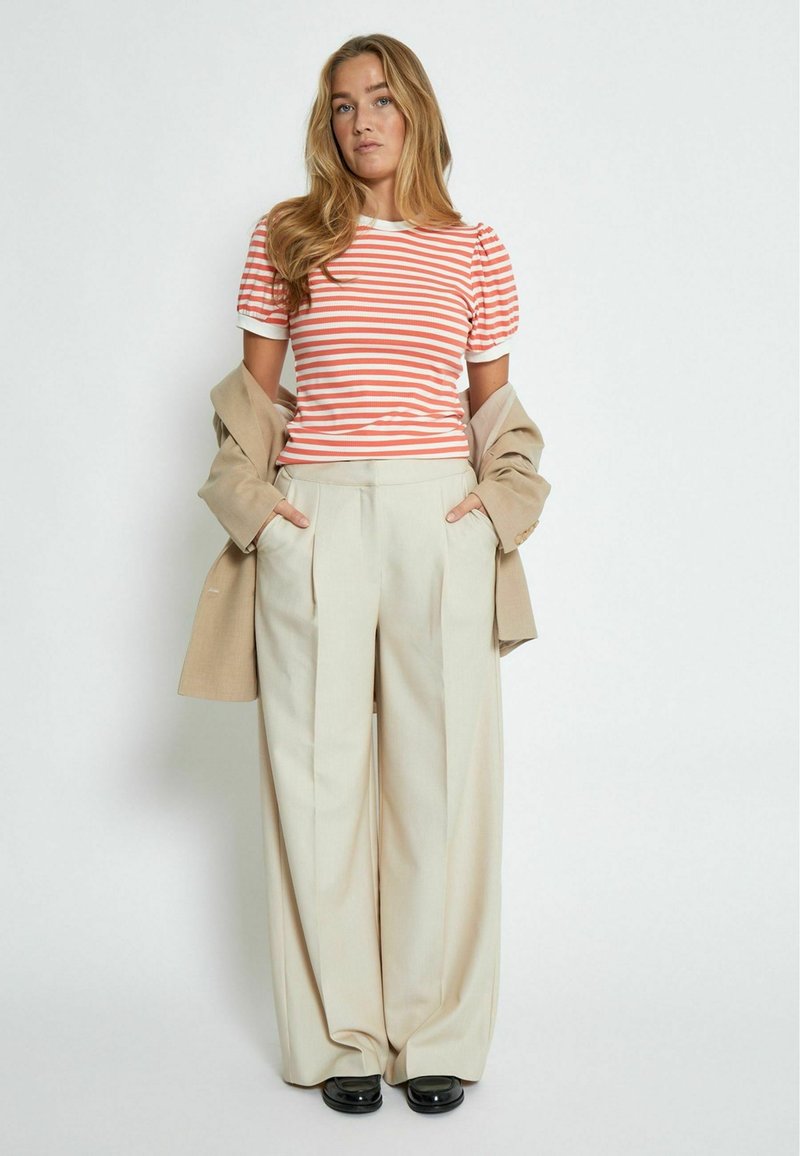 T-shirt a righe arancione e bianco con maniche a sbuffo, abbinato a pantaloni beige a gamba larga. Un blazer beige è adagiato sulle spalle.